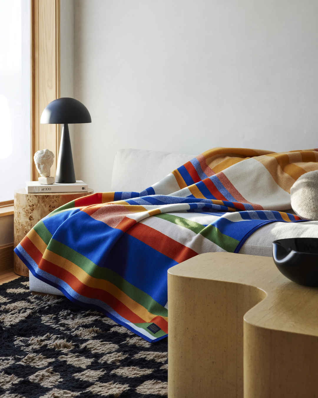 Colorful striped blanket draped on a white surface, accompanied by a black mushroom lamp, a stack of books, and light wood furniture.