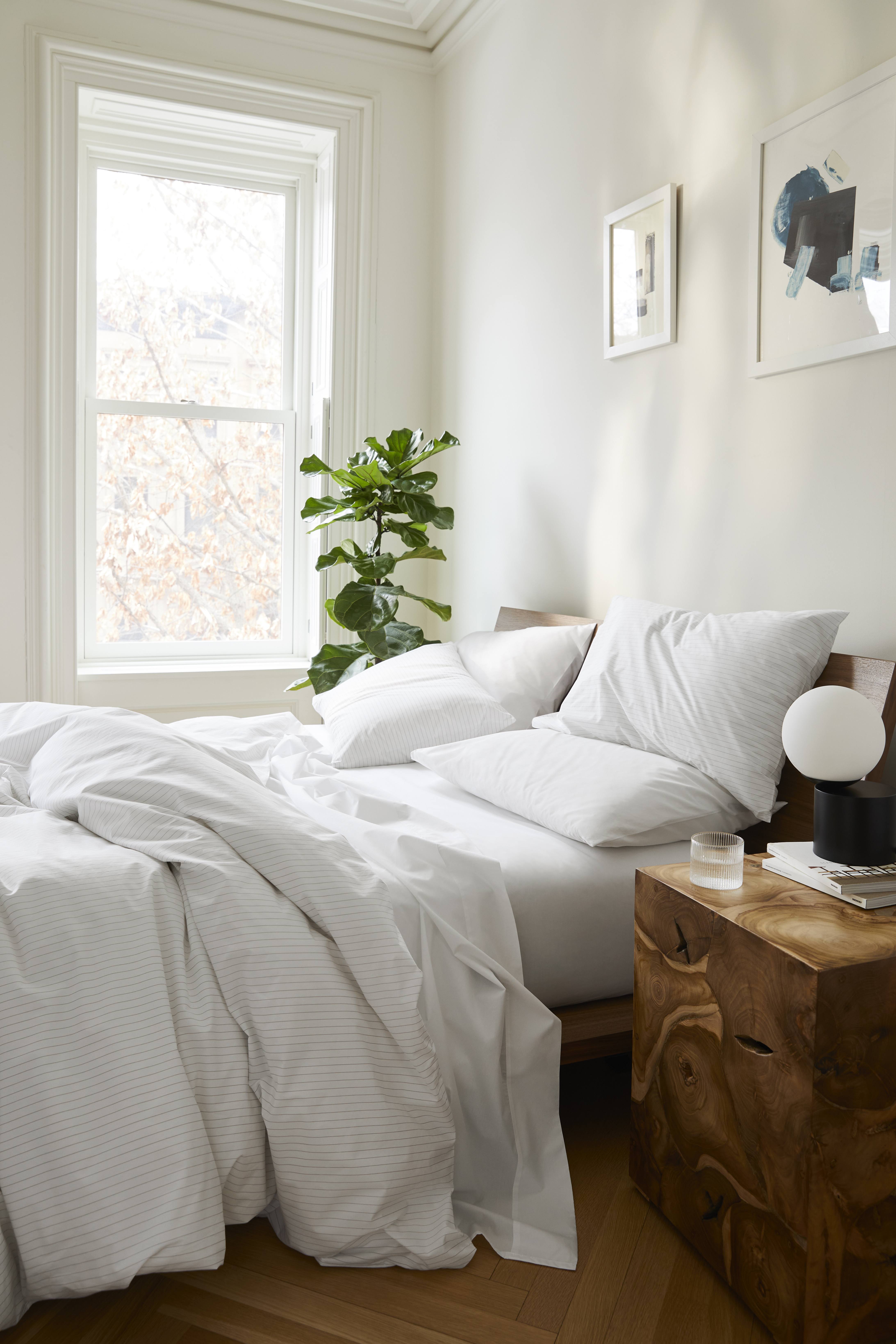Bright bedroom interior with a bed dressed in white striped bedding next to a wooden nightstand, illuminated by natural light.