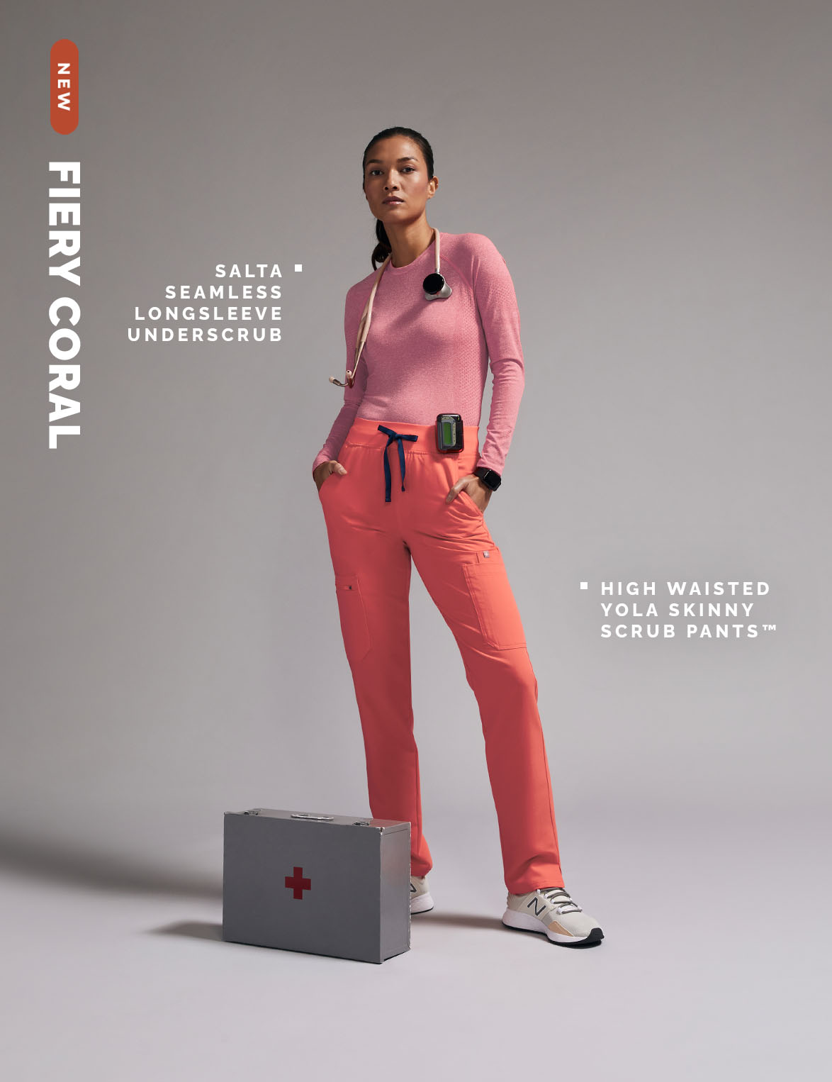 Asian female nurse in coral scrubs and long-sleeved shirt poses indoors near a gray first aid box for a healthcare fashion shoot.