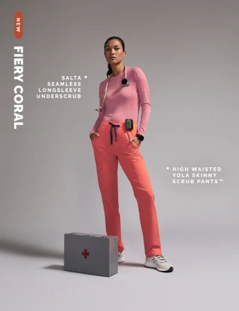 Asian female nurse in coral scrubs and long-sleeved shirt poses indoors near a gray first aid box for a healthcare fashion shoot.