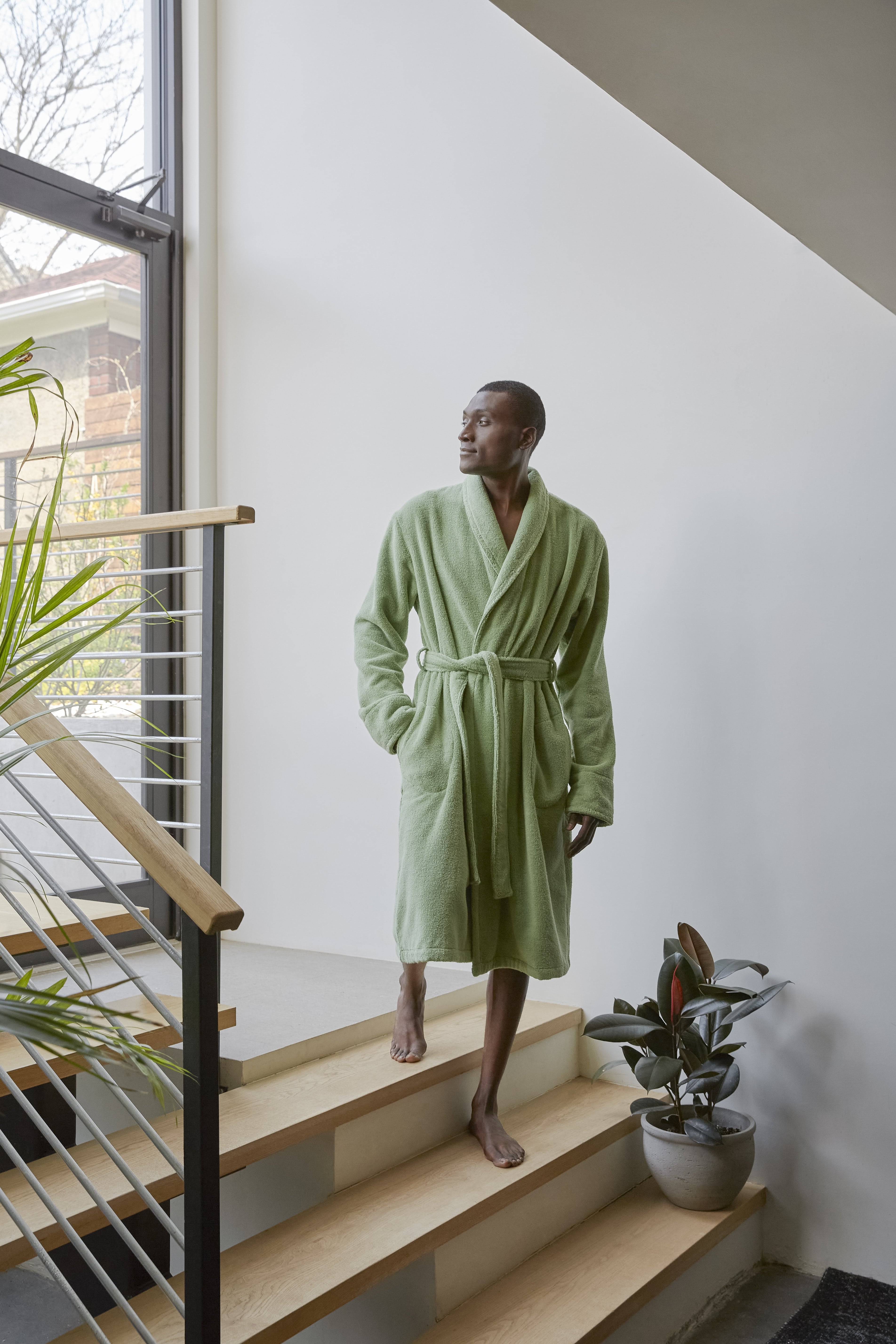 An African American man walks down wooden stairs in a light green bathrobe in a modern home with potted plants and a large window.