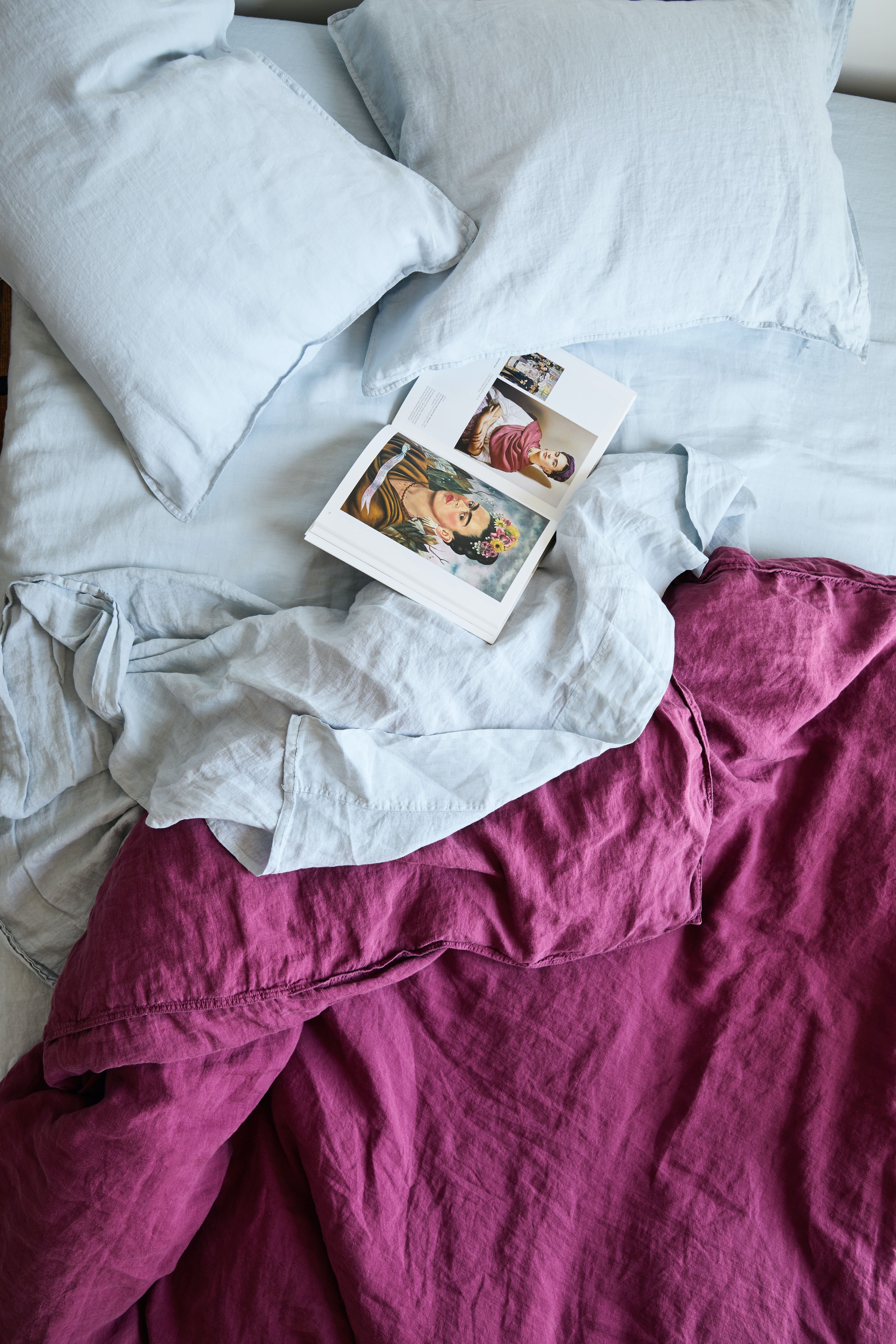 Overhead shot of an unmade bed with blue sheets, a dark pink blanket, two pillows, and an open book featuring artwork.