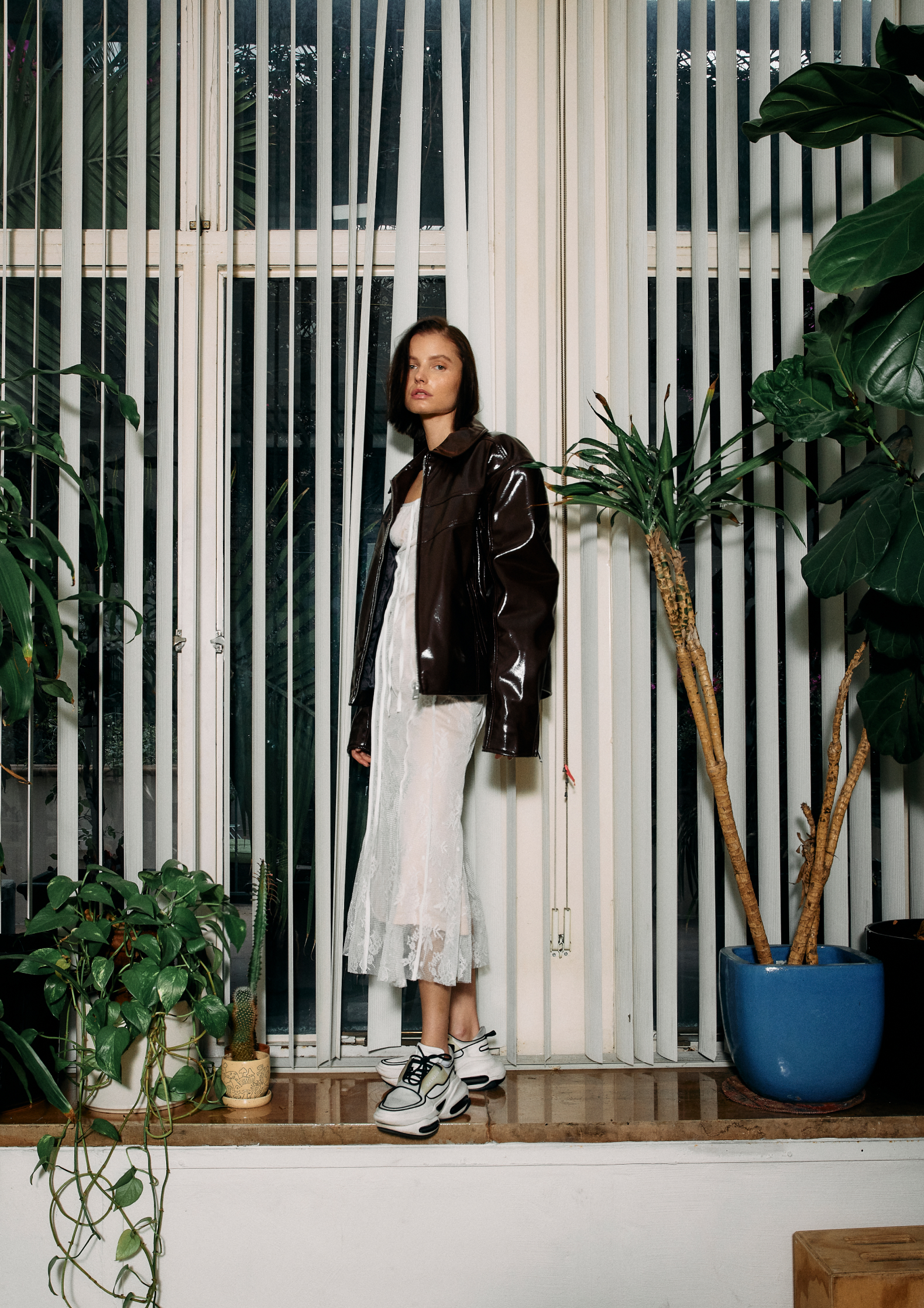 A woman stands in front of a window with vertical blinds wearing a brown jacket, white dress, and sneakers, surrounded by potted plants.