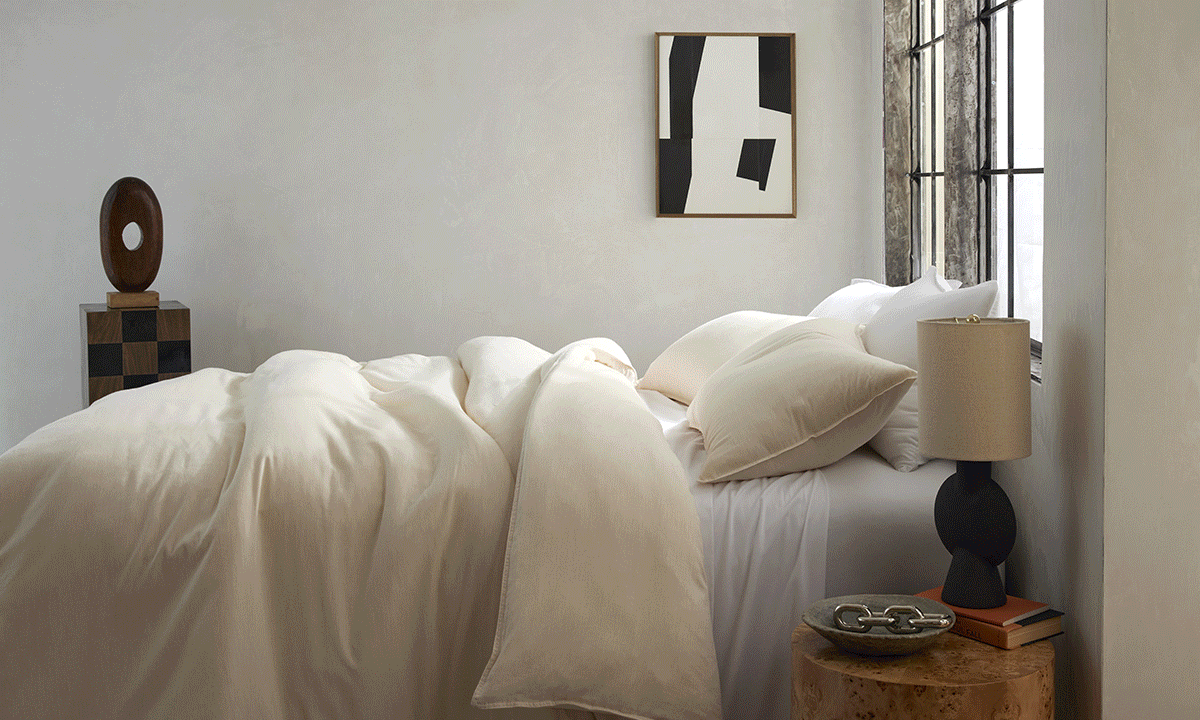 Bedroom interior featuring a cream-colored duvet and pillows on a bed, a decorative side table with a lamp, and abstract art on the wall.