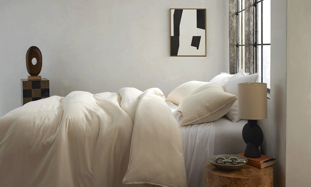 Bedroom interior featuring a cream-colored duvet and pillows on a bed, a decorative side table with a lamp, and abstract art on the wall.