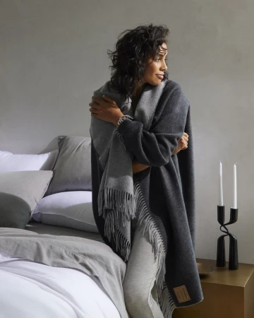 Elegant woman wrapped in a fringed grey and charcoal blanket, standing near a bed with soft pillows and a black candlestick.