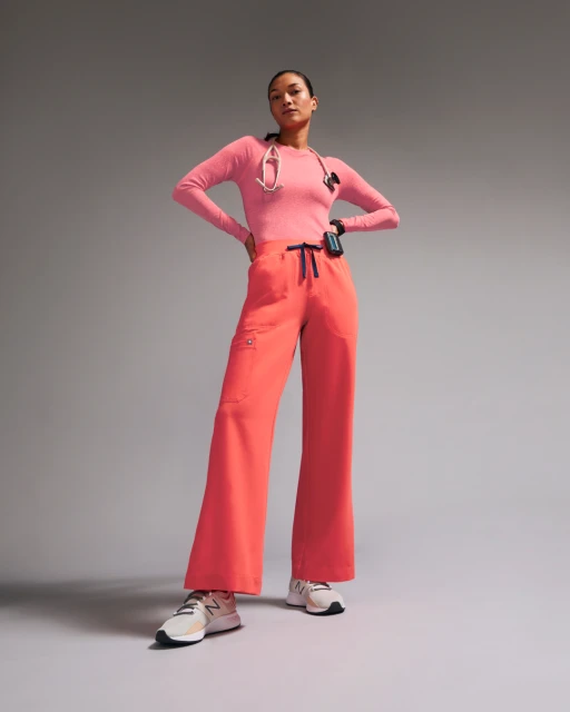 Female healthcare worker stands in a pink long-sleeve shirt and coral wide-leg pants with stethoscope and sneakers in a photo studio.