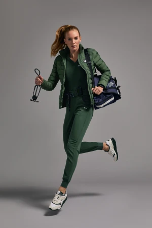 A woman wearing green scrubs and a puffy coat sprints in a studio holding a stethoscope and gym bag, showcasing healthcare fashion.