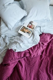 An overhead view of a bed with light blue linen pillows, a light blue duvet, an open book, and a folded magenta linen blanket.
