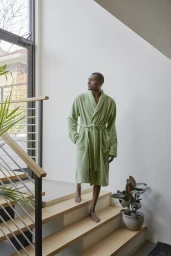 Man wearing a green bathrobe walking down indoor wooden stairs by a large window with plants