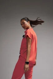 Woman in vibrant coral medical scrubs and a stethoscope turns her head toward the camera against a grey seamless backdrop.