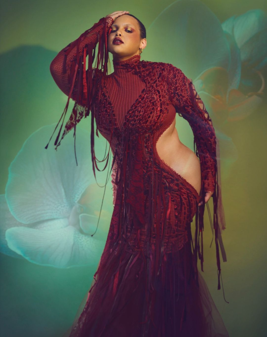 Elegant adult female fashion model poses in a textured maroon dress with fringe against a soft green floral backdrop in a studio setting.