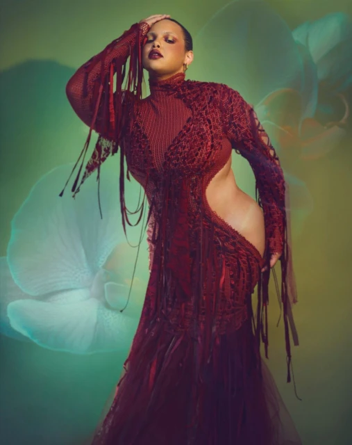 Elegant adult female fashion model poses in a textured maroon dress with fringe against a soft green floral backdrop in a studio setting.