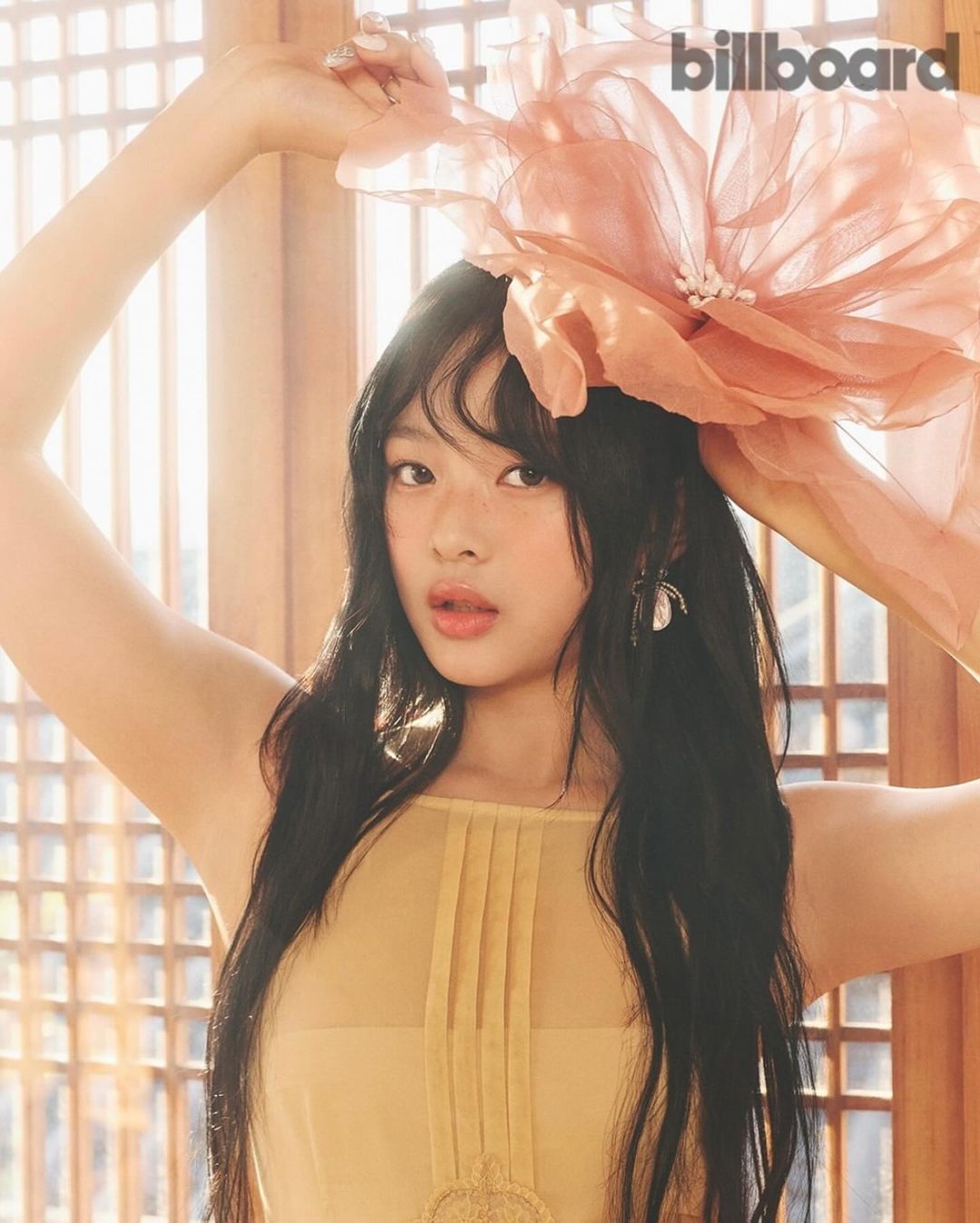 Indoor portrait of a woman with long dark hair, wearing a sheer yellow top and a large pink flower headpiece, with billboard logo.