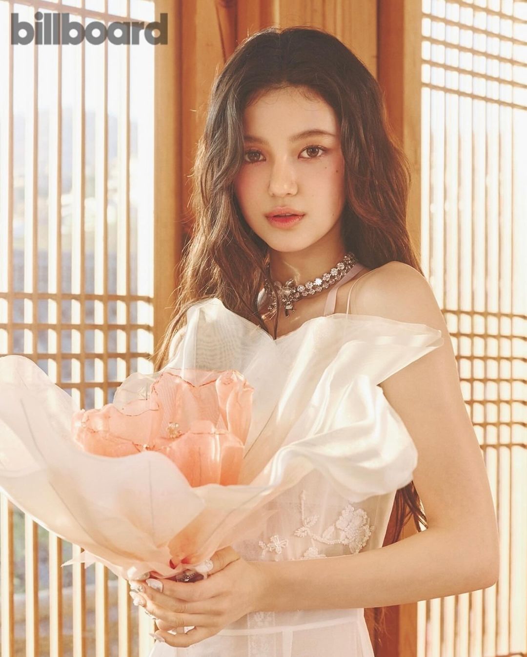 Portrait of a young woman holding a bouquet of light orange tulips, adorned with a sparkling necklace, in a room with traditional window panes.