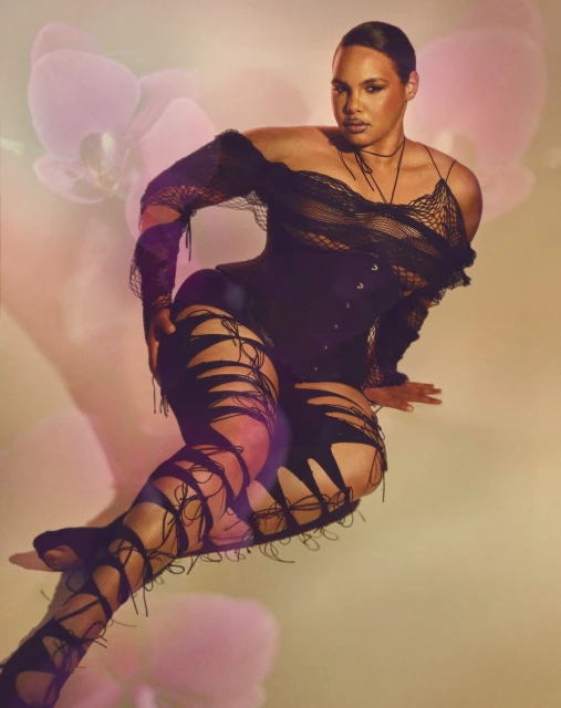 Woman posing in black off-the-shoulder top, corset, and thigh-high ripped leggings with orchid flower background in a studio setting.
