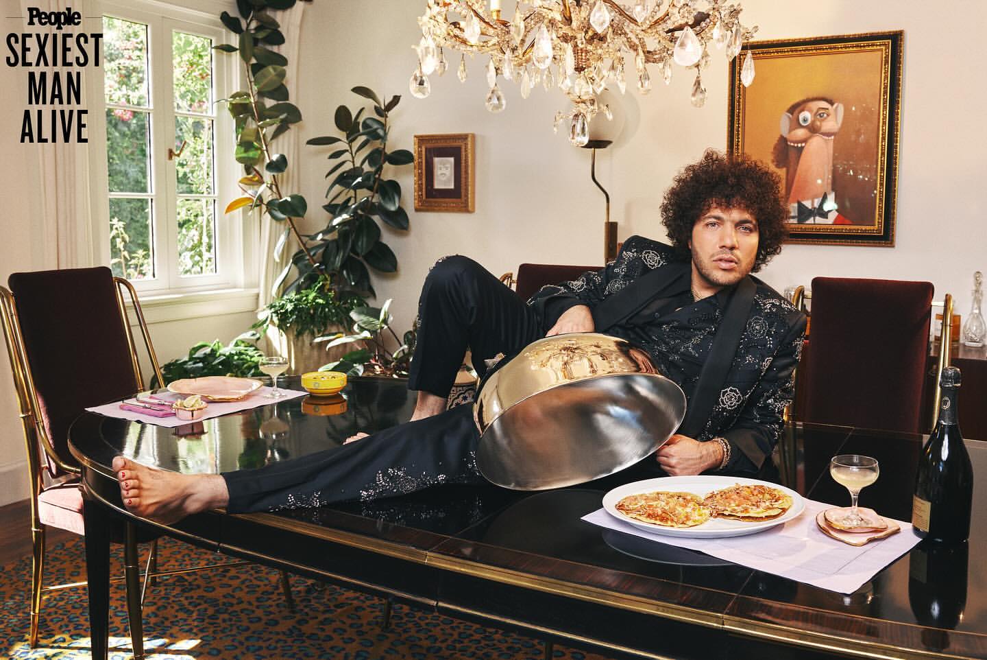 Adult male with afro, dressed in black floral suit, reclines on black dining table with food, plants, and chandelier in interior setting.
