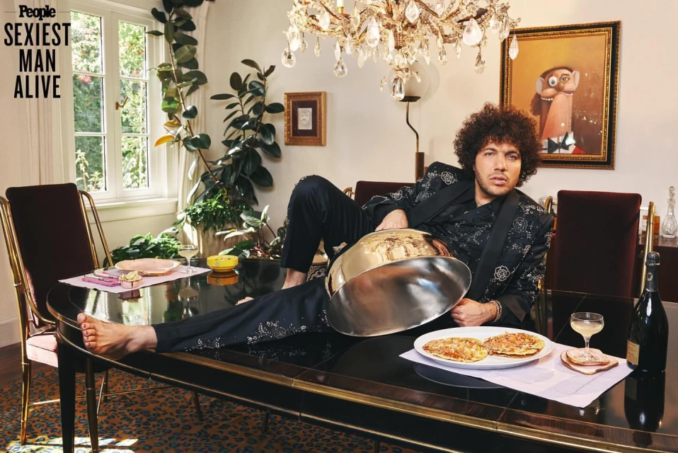 Adult male with afro, dressed in black floral suit, reclines on black dining table with food, plants, and chandelier in interior setting.
