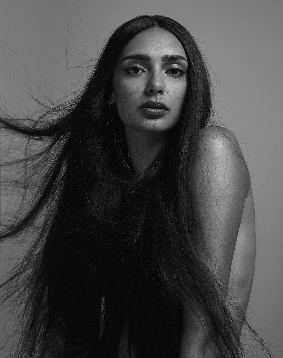 Black and white portrait of a woman with long dark hair flowing around her face, looking directly at the camera against a grey backdrop.