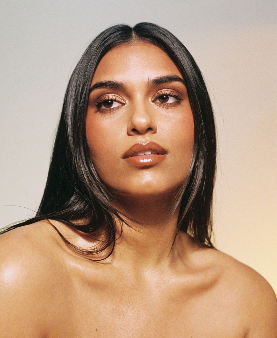 Close-up portrait of a young adult woman with long, dark hair, fair skin, and glossy lips, looking off to the side against a gradient background.