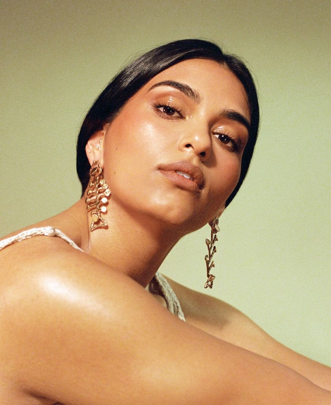Woman with dark hair poses wearing large gold earrings against a light green background in a beauty or fashion portrait.