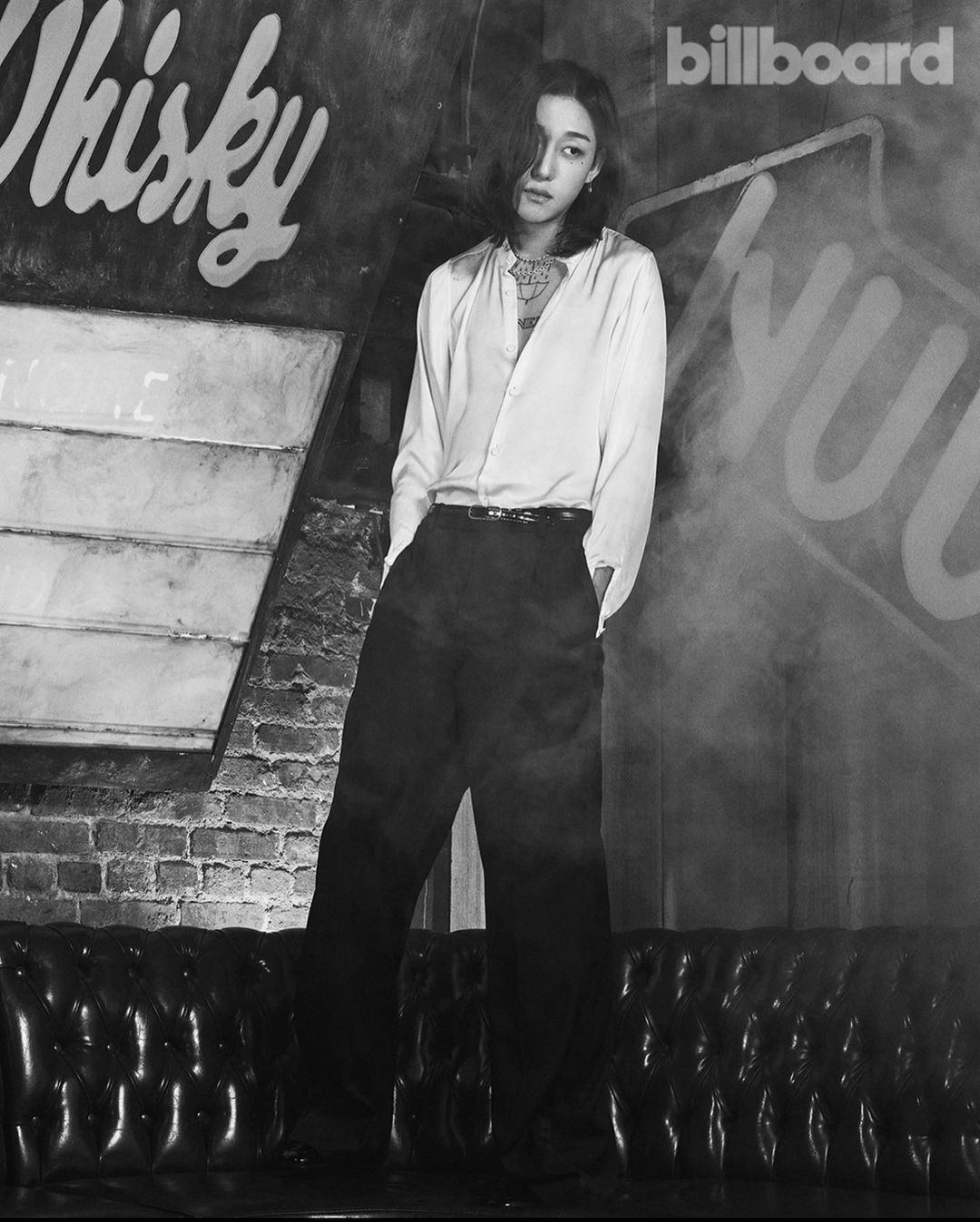 Monochrome portrait of a slender adult in an unbuttoned white shirt and dark trousers standing in front of a graffiti wall.