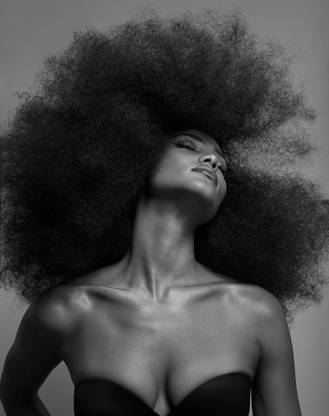 Monochrome portrait of a Black woman with a large afro hairstyle, eyes closed, wearing a black strapless top, against a gray background.