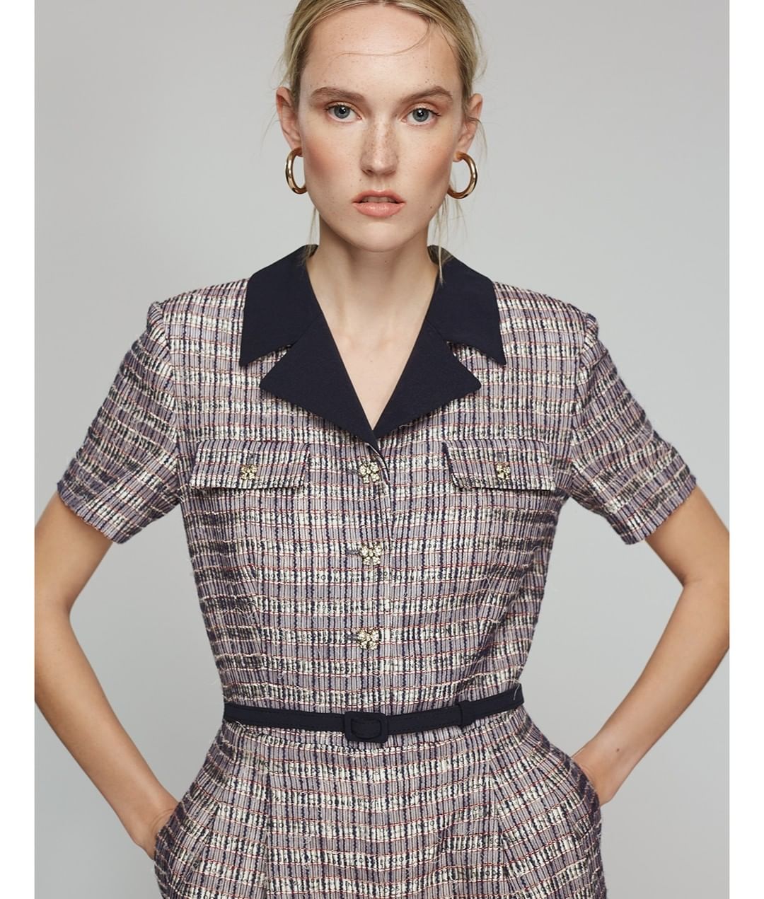Woman wearing a tweed dress with navy blue collar and gold hoop earrings in a studio portrait with a light gray background.