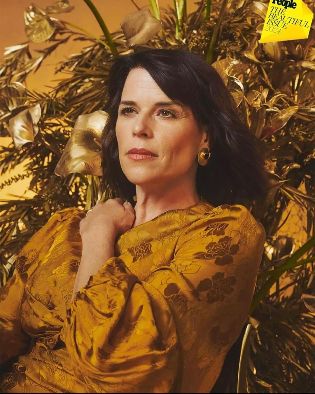 Close-up portrait of Neve Campbell in People's 'The Beautiful Issue' 2024, in a golden floral dress against a gilded floral background.
