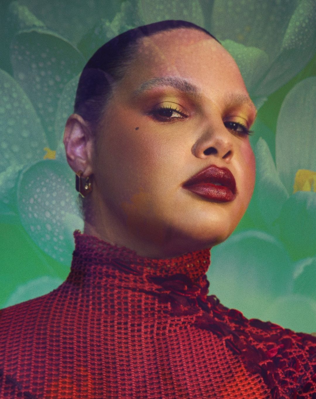 Close-up studio portrait of a woman with dark lipstick and creative makeup, set against a backdrop of large green and white flowers.
