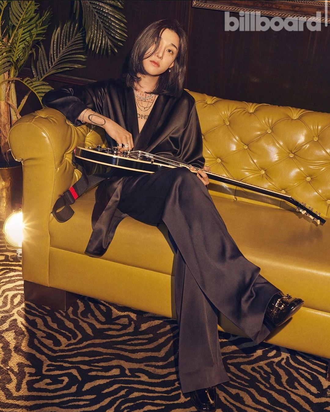 A person sits on a yellow tufted couch while holding a guitar, in a dimly lit room with a plant and a Billboard logo in the shot.