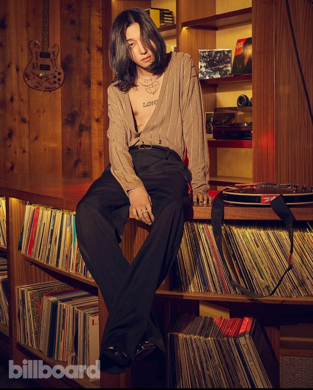 Stylish person with long hair poses in a warm-toned photo studio setting, wearing a cardigan, dress pants, and rings, surrounded by vinyl records.