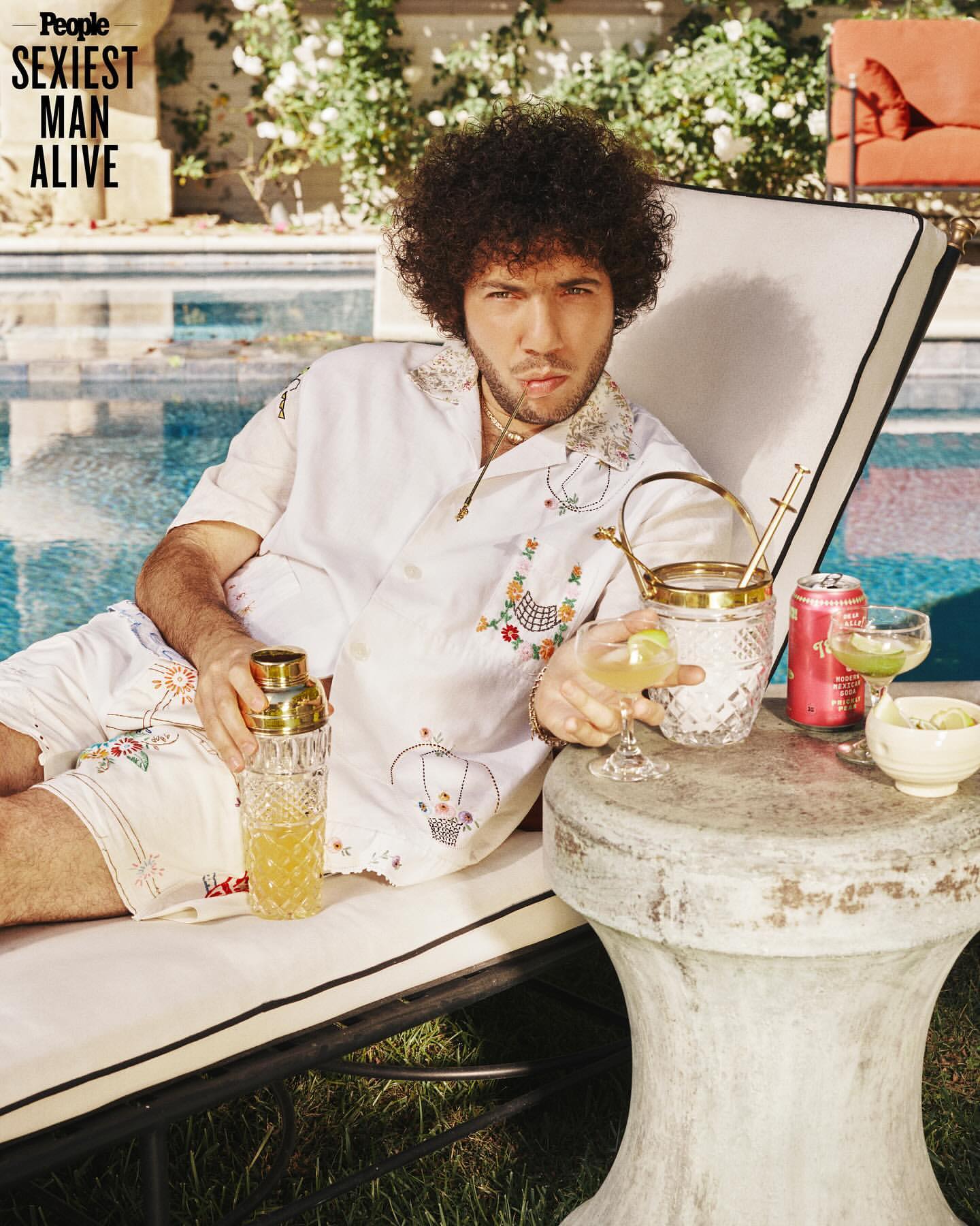 Man with afro lounging by a pool holding a cocktail shaker, wearing a patterned white shirt and shorts with drinks and an ice bucket.