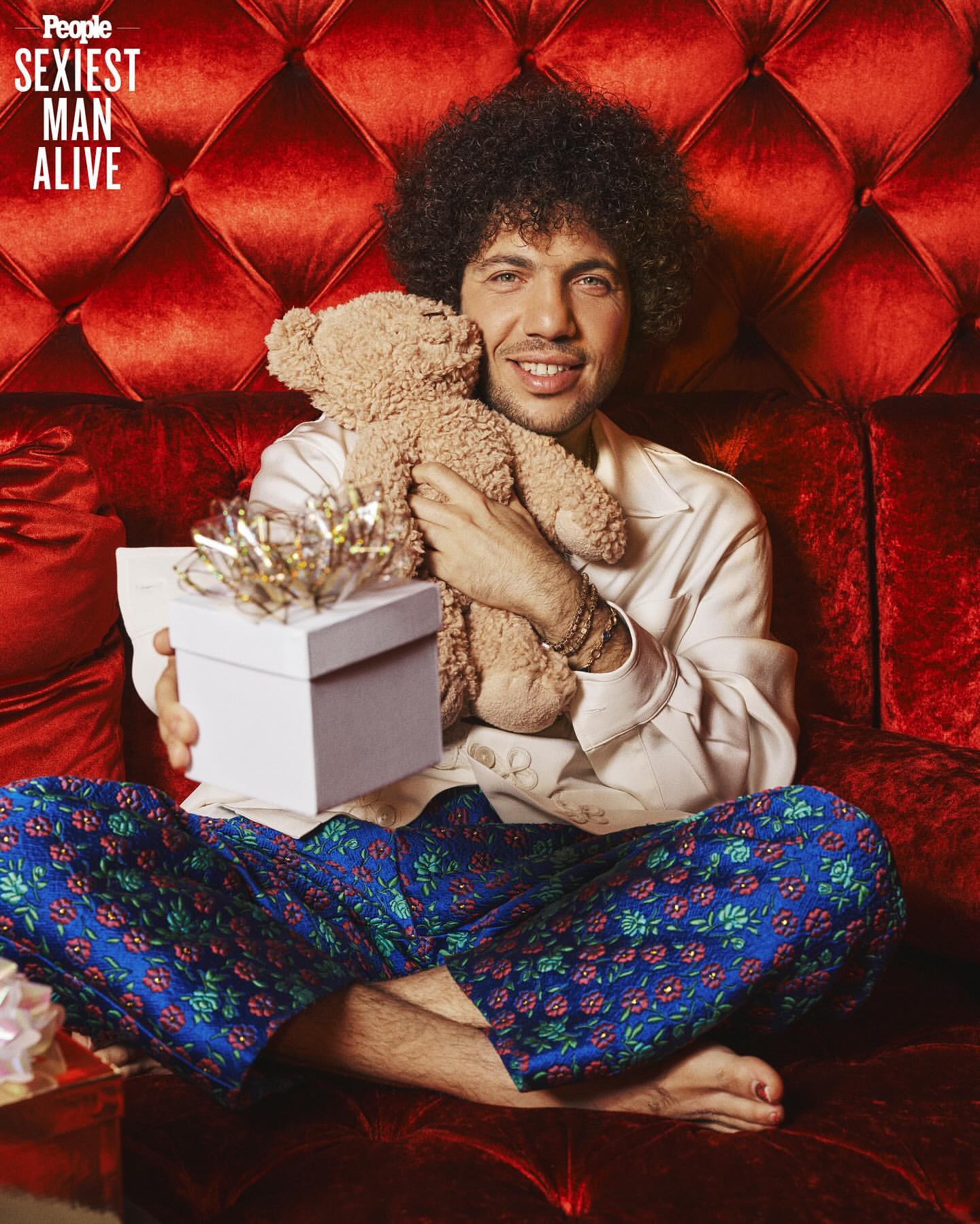Adult male poses on red velvet couch with teddy bear and gift box, wearing patterned pajama pants in indoor setting with bright lighting.