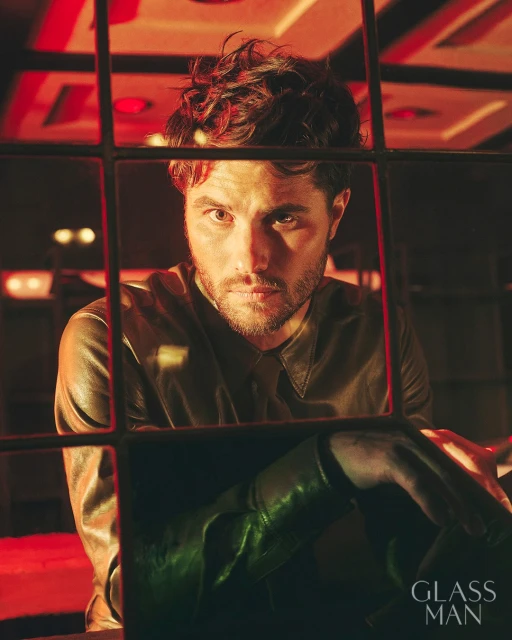 Portrait of a male in a leather jacket with dark hair behind a window with bars, featuring moody red lighting and a shadowed foreground.