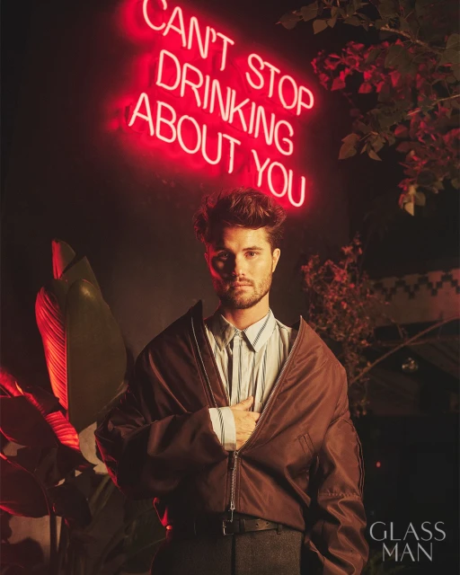 Portrait of a handsome adult male model in a brown bomber jacket, posing in front of a neon sign and greenery.