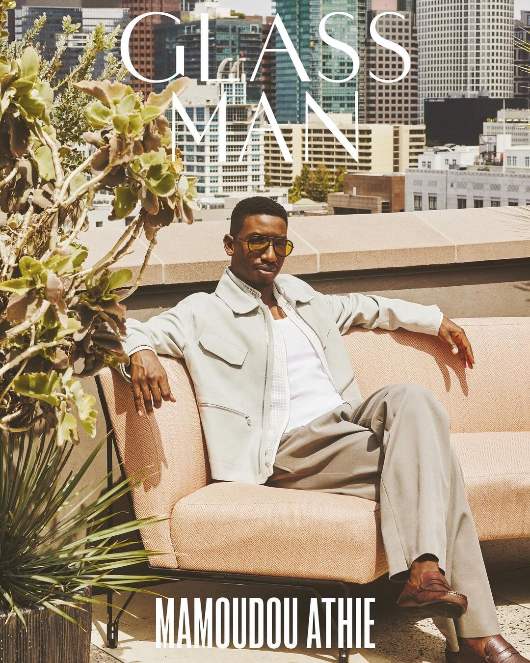 Male model Mamoudou Athie in neutral tones on a peach sofa, rooftop city view. Fashion editorial, modern style, urban setting, stylish portrait.