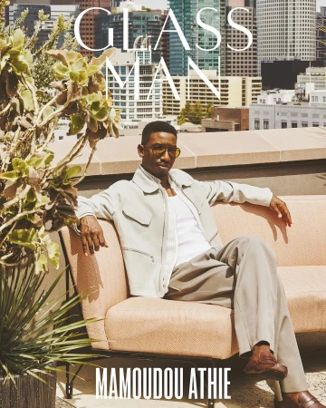 Male model Mamoudou Athie in neutral tones on a peach sofa, rooftop city view. Fashion editorial, modern style, urban setting, stylish portrait.
