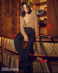 Man with long dark hair and tattoos wearing striped top and dark trousers seated in a record store