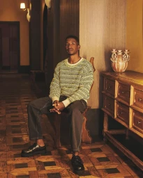 Black man seated on a wooden chair in an interior hallway wearing a patterned sweater and dark trousers