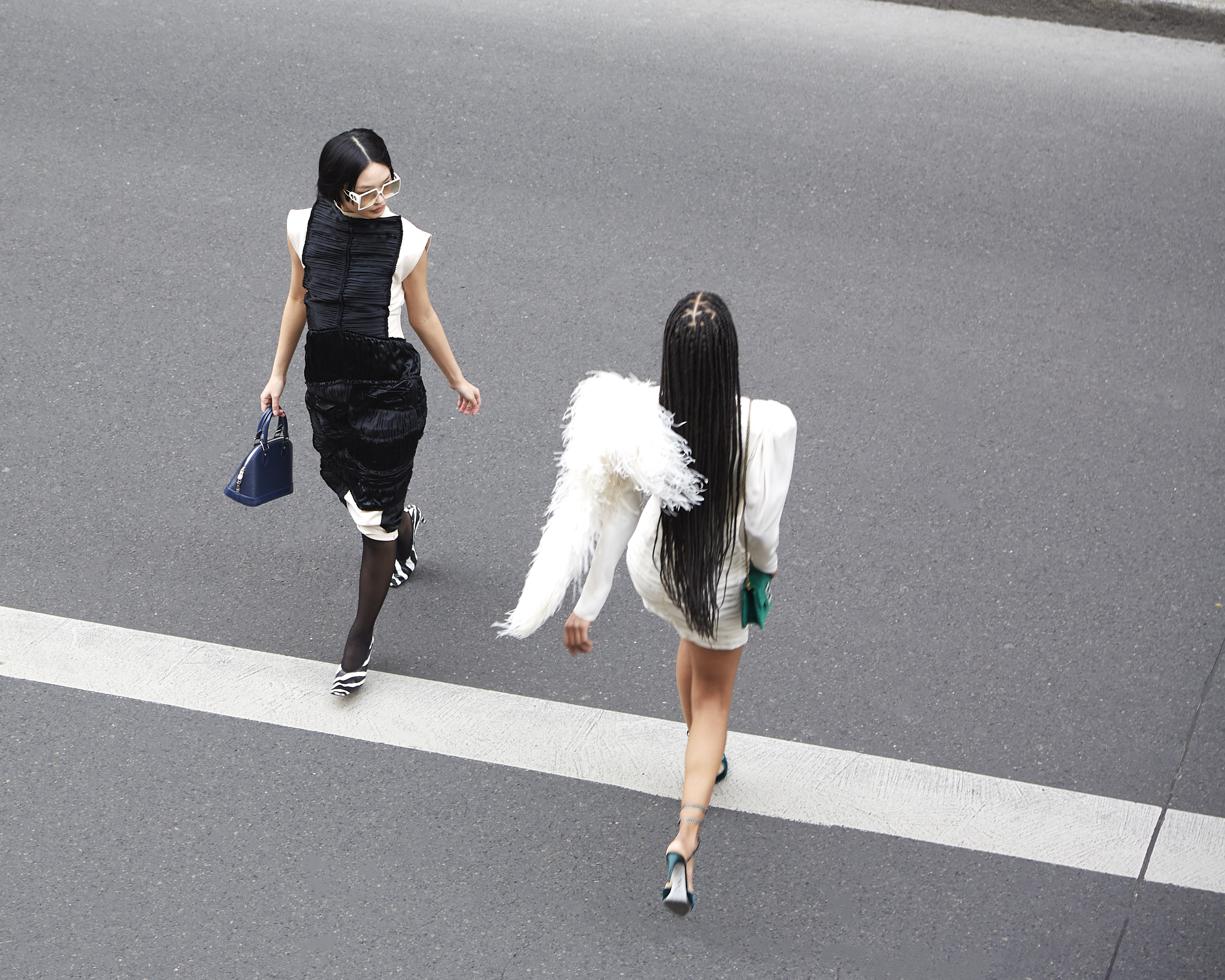 Two women in fashionable dresses walk across a crosswalk with purses. One woman has braids and feathered shoulder accents.