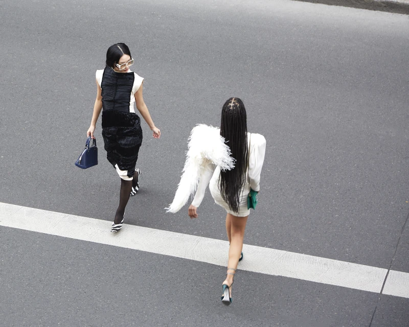 Two women in fashionable dresses walk across a crosswalk with purses. One woman has braids and feathered shoulder accents.