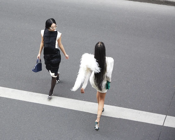 Two women in fashionable dresses walk across a crosswalk with purses. One woman has braids and feathered shoulder accents.