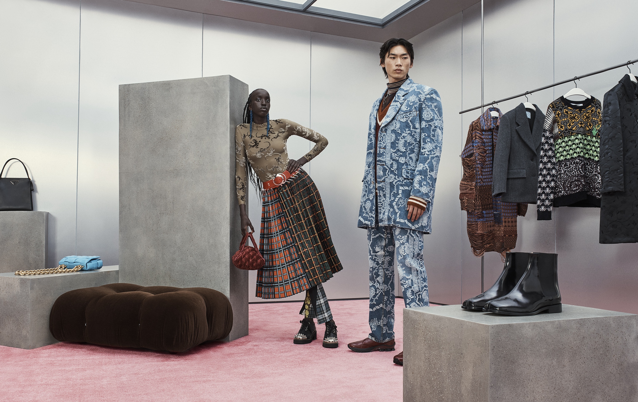 Two fashion models pose indoors on a pink carpet near clothing rack and display cases. The models wear patterned suit and skirt outfits.