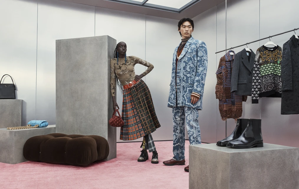 Two fashion models pose indoors on a pink carpet near clothing rack and display cases. The models wear patterned suit and skirt outfits.