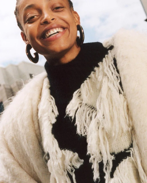 Smiling woman with short hair and hoop earrings poses wearing a black turtleneck sweater and fuzzy white cardigan with fringe.