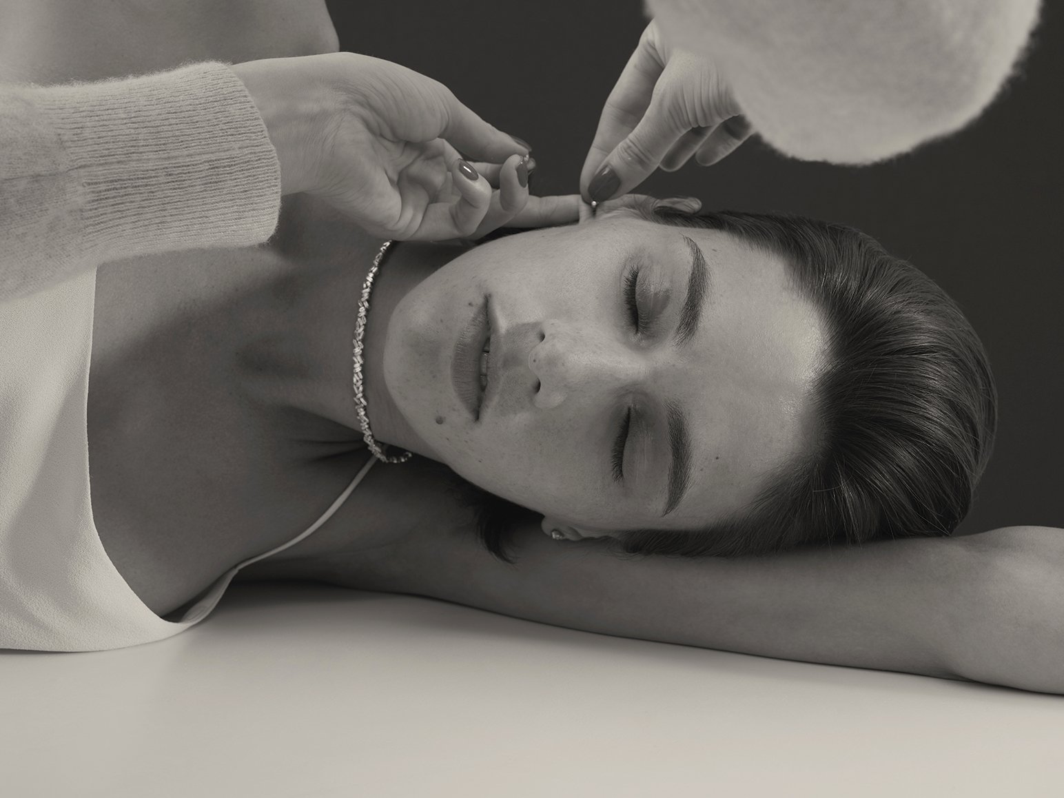 Woman reclines as hands adjust her earring in a close-up studio shot showcasing beauty and subtle glamour, set against a dark background.