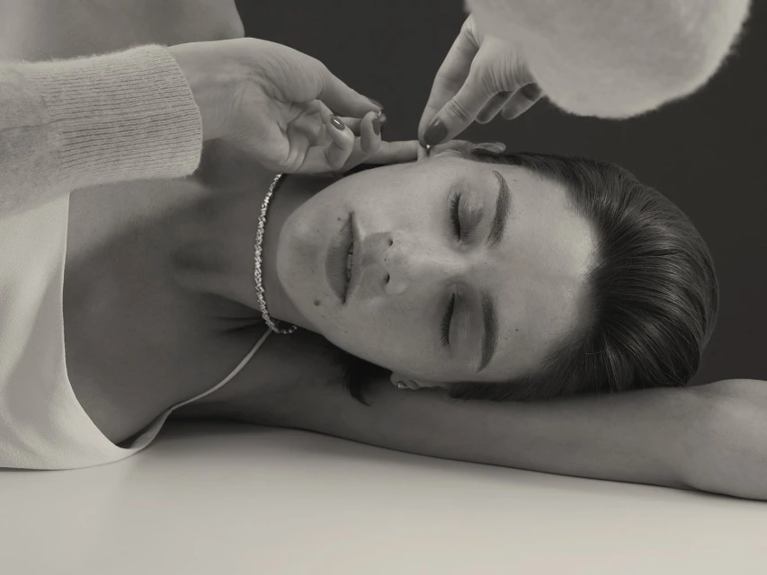 Woman reclines as hands adjust her earring in a close-up studio shot showcasing beauty and subtle glamour, set against a dark background.