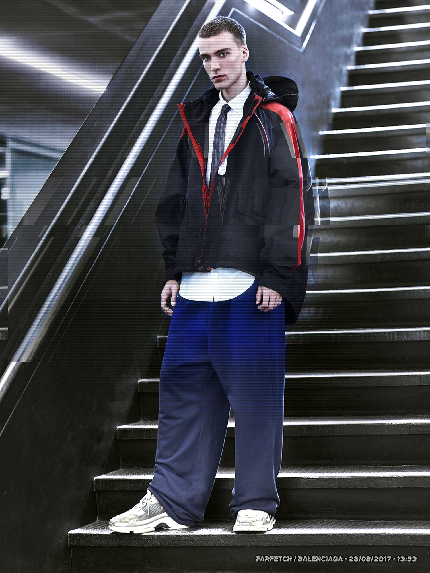 A young adult man poses on stairs in a black hooded jacket with red trim, a white shirt, tie, and blue pants, in an indoor setting.