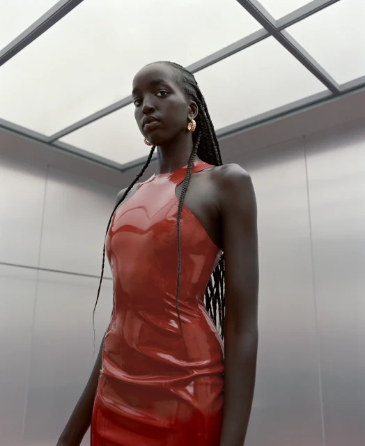 A fashionable Black woman in a red latex dress poses in an elevator with neutral background. She has long braids and is wearing gold earrings.